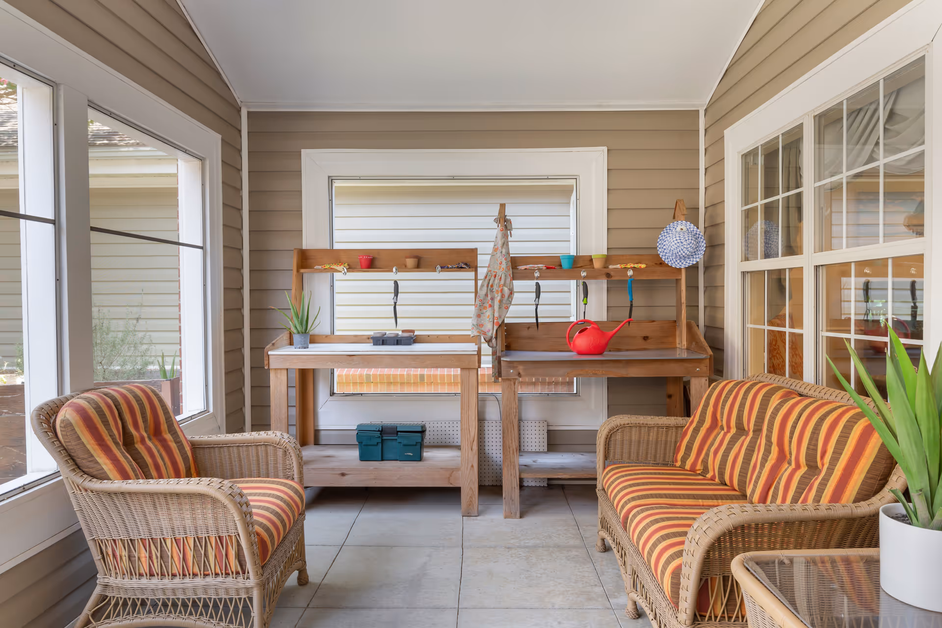A cozy indoor porch area with wicker furniture including a striped cushioned armchair and loveseat. There are two wooden potting benches against the wall with gardening tools, small pots, a red watering can, and a hanging apron. Large windows surround the space, letting in natural light.