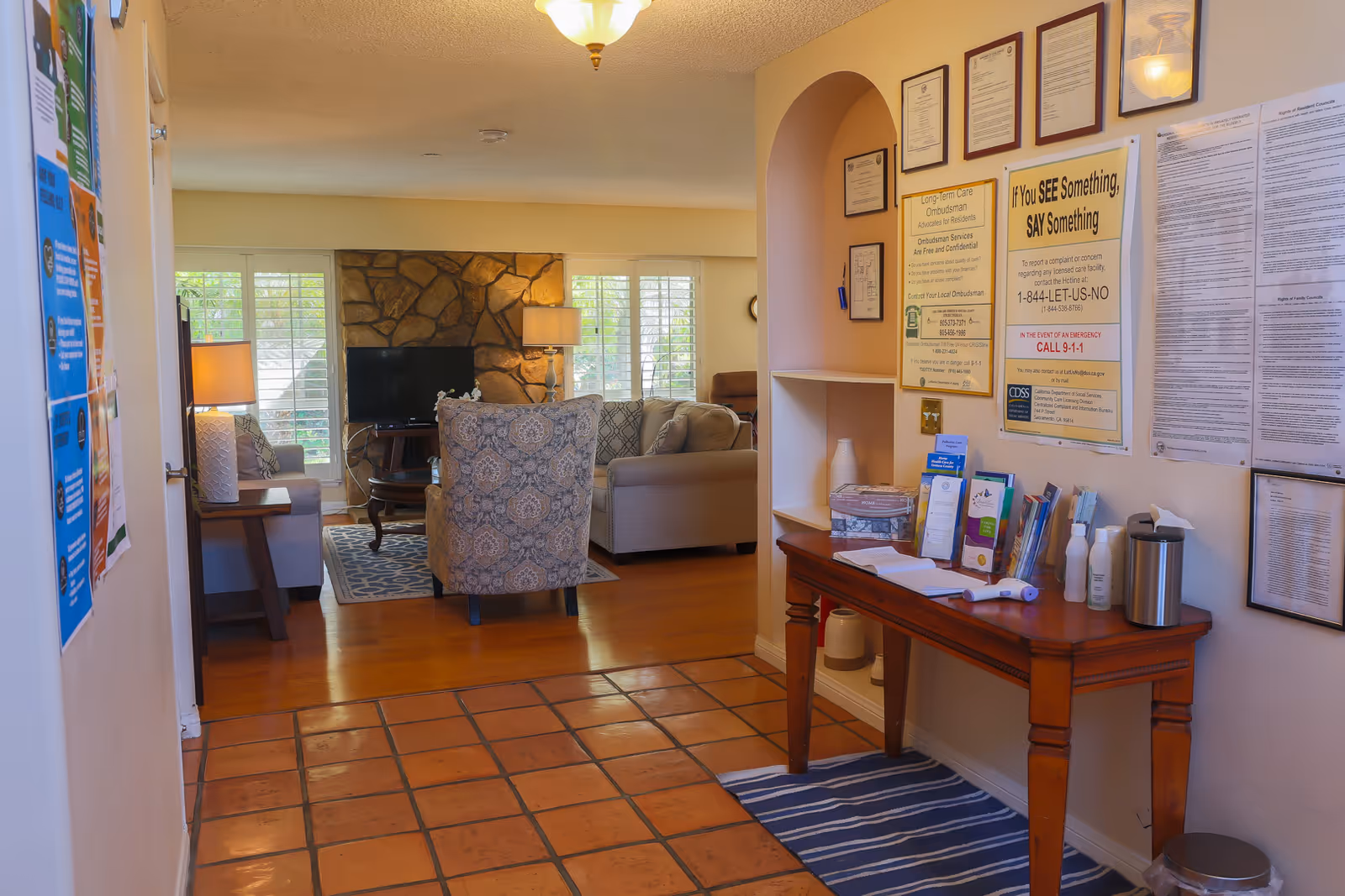 Interior view of a senior living facility showing a hallway with informational posters and brochures on the right wall and a wooden table with various pamphlets and items. The hallway leads into a cozy living room area with a patterned armchair, sofas, a coffee table, a TV mounted on a stone fireplace, and windows with white shutters letting in natural light.