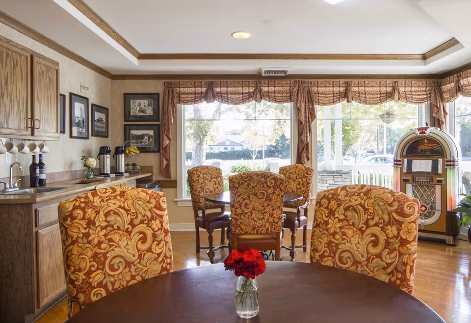 A cozy dining area in a senior living facility with round wooden tables and upholstered chairs featuring a red and gold floral pattern. A small vase with red flowers is on the closest table. The room has large windows with patterned valances letting in natural light. To the left, there is a countertop with a sink, coffee dispensers, and framed black and white photos on the wall. On the right side of the room, there is a vintage-style jukebox.