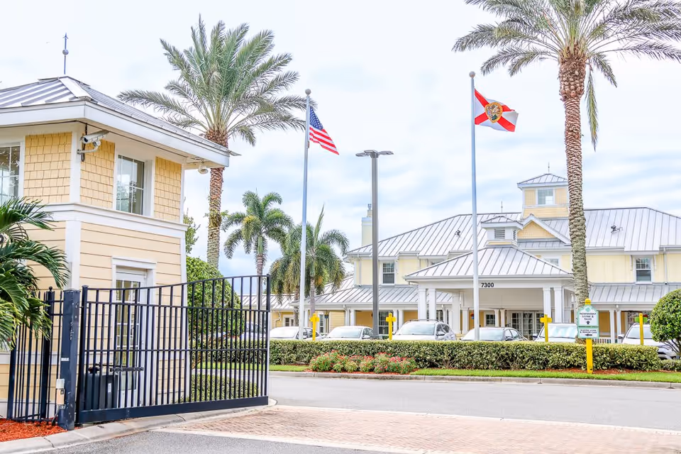 Entrance gate and guardhouse of The Brennity at Melbourne Senior Living facility with palm trees, American and Florida state flags, and a large building with a metal roof in the background.