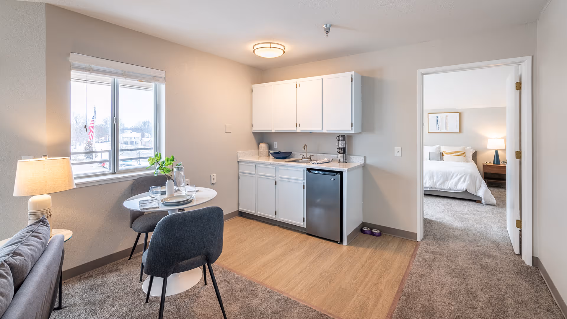 A small apartment interior featuring a kitchenette with white cabinets, a mini refrigerator, and a sink. Adjacent to the kitchenette is a round dining table set for two with plates, glasses, and a small plant. A window with blinds lets in natural light. To the right, an open doorway leads to a bedroom with a bed, nightstand, and lamp. The flooring is a combination of carpet and wood-like material.