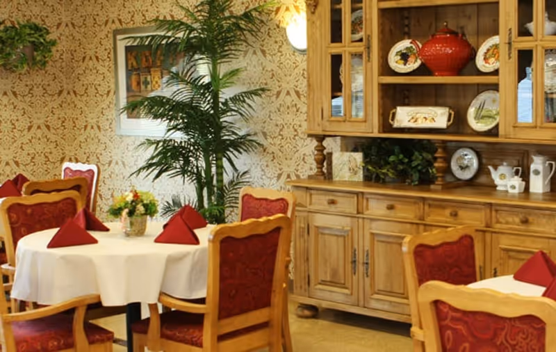 A cozy dining room with round tables covered in white tablecloths, each set with red folded napkins and a small floral centerpiece. The room features wooden chairs with red upholstered seats and backs, a wooden cabinet displaying decorative plates and a red teapot, and a tall green plant against a patterned wallpapered wall.