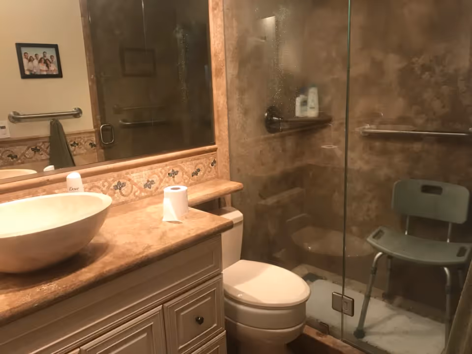 Bathroom with a beige countertop and a round vessel sink, a toilet, and a glass-enclosed shower area with a green shower chair and grab bars. A large mirror is above the sink, and a framed photo hangs on the wall.