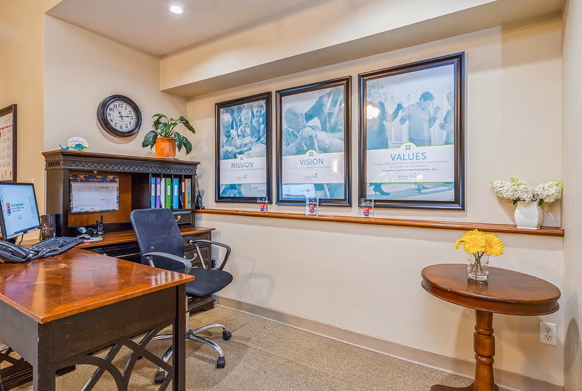 An office area with a wooden desk and black office chair. On the desk are a computer monitor, keyboard, phone, and some office supplies. Behind the desk is a wooden hutch with binders and a potted plant. On the wall above the hutch are three framed posters labeled Mission, Vision, and Values. To the right, a small round wooden table holds a vase with yellow flowers, and a white vase with white flowers is on a ledge. A clock is mounted on the wall above the hutch.