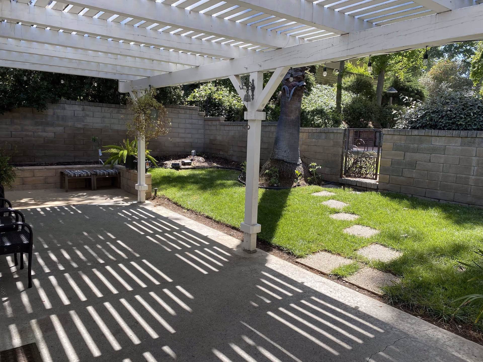 Outdoor patio area with a white pergola casting striped shadows on the concrete floor. There are black chairs on the left side and a small bench with cushions against a brick wall. A green lawn with stepping stones leads to a decorative metal gate in the brick wall. Trees and bushes surround the area providing greenery and shade.