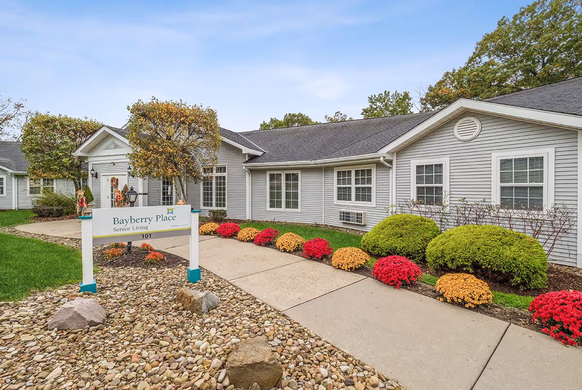 Exterior view of Bayberry Place Senior Living facility showing a single-story building with gray siding, white trim, and multiple windows. There is a concrete walkway lined with red and yellow flowers and green shrubs leading to the entrance. A sign in front reads 'Bayberry Place Senior Living 101'. The sky is clear with some light clouds.