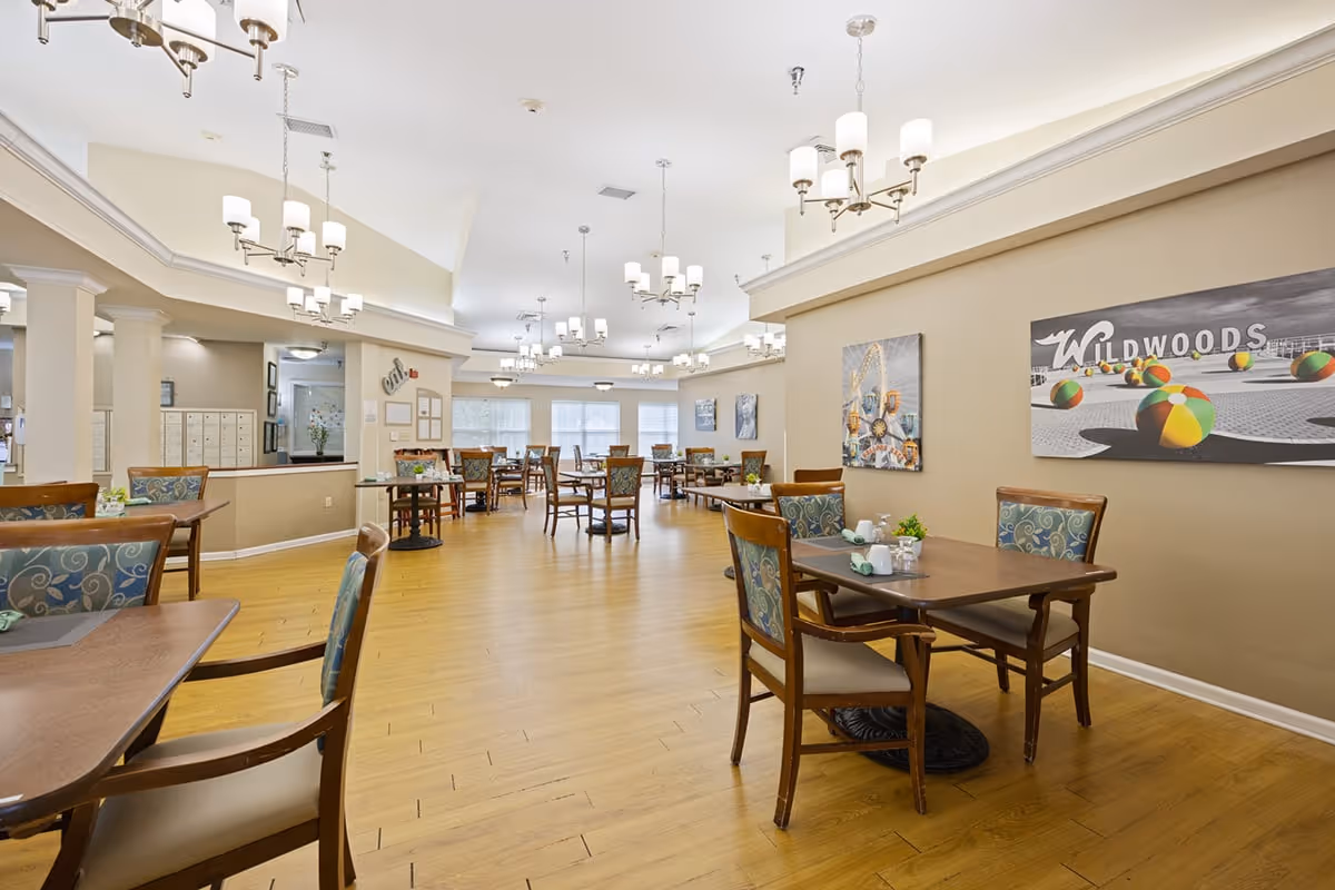 A spacious dining room in a senior living facility with multiple wooden tables and chairs arranged neatly. The room features light-colored walls, wooden flooring, and several chandeliers hanging from the ceiling. Wall art includes a large picture with the word 'WILDWOODS' and colorful beach balls. The area is well-lit with natural light coming through large windows in the background.