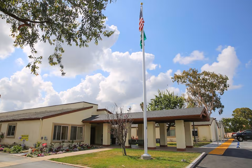 Exterior view of Glenwood Care Center building with a covered entrance, an American flag and another flag on a flagpole, a small garden with flowers, and a parking area under a partly cloudy sky.