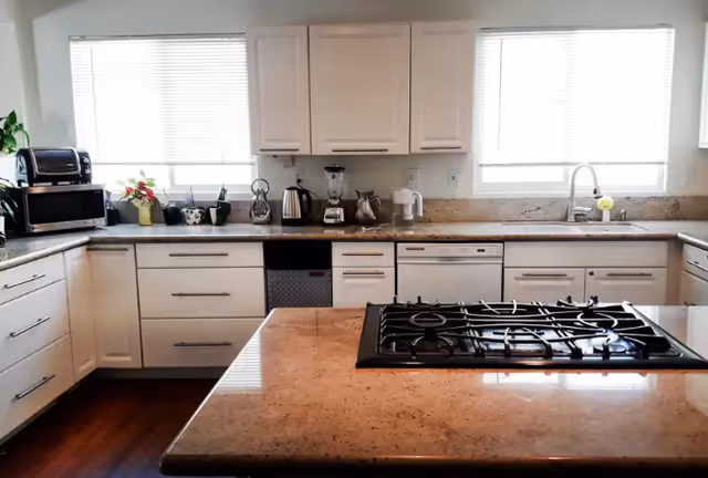 Bright modern kitchen with a central island gas cooktop, white cabinets, countertop appliances, and a sink under windows.
