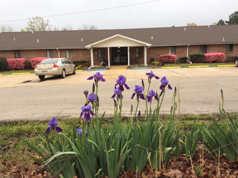 Exterior view of a single-story brick building with a covered entrance and a parking lot in front. Purple irises and green plants are in the foreground, with some cars parked near the entrance. The sky is overcast.