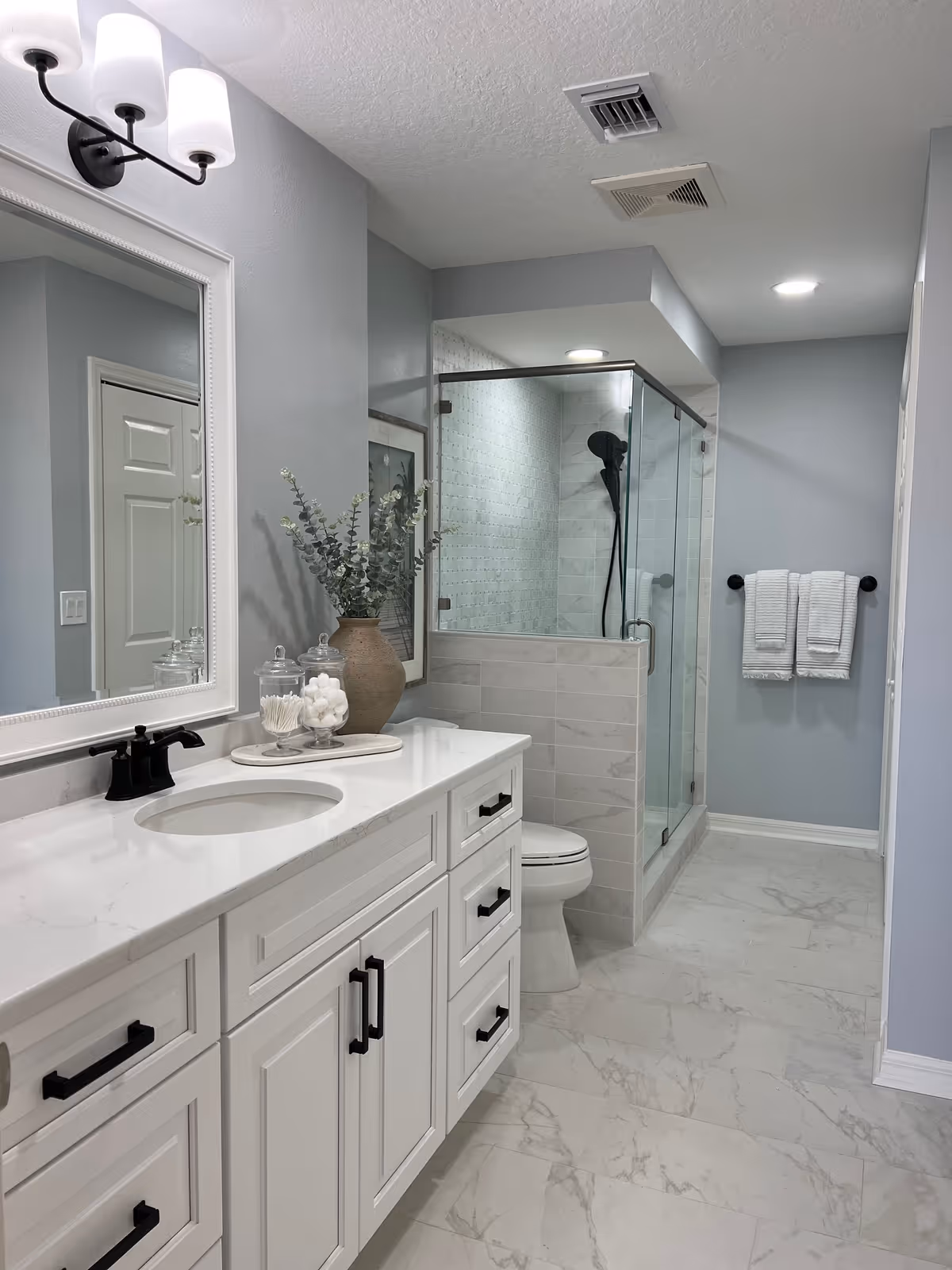 Bright modern bathroom with a white vanity and marble countertop, glass-enclosed shower, toilet, and towel rack.