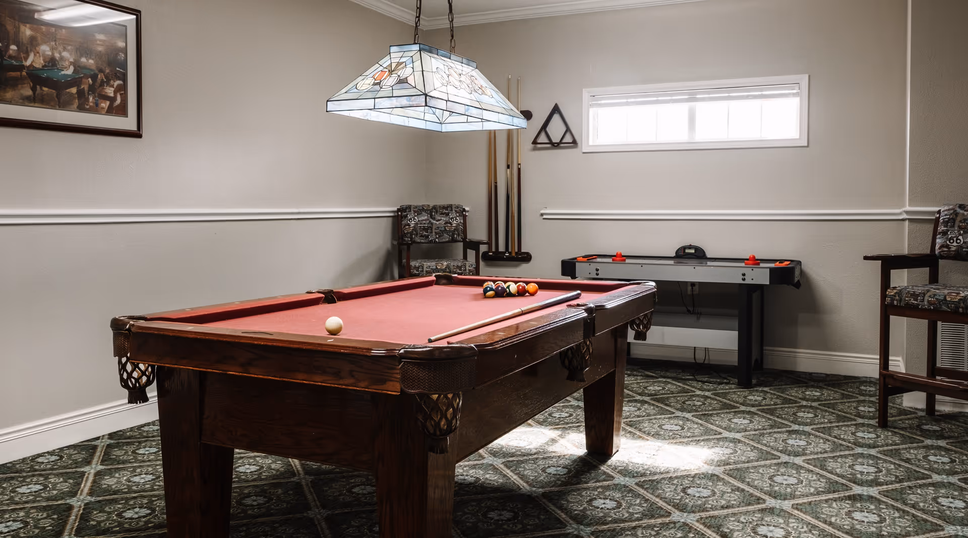 A game room with a red felt pool table in the center, pool cues on the table, and a hanging stained glass light fixture above. There is an air hockey table against the far wall, two chairs with patterned upholstery, a small window, and a framed picture on the wall.