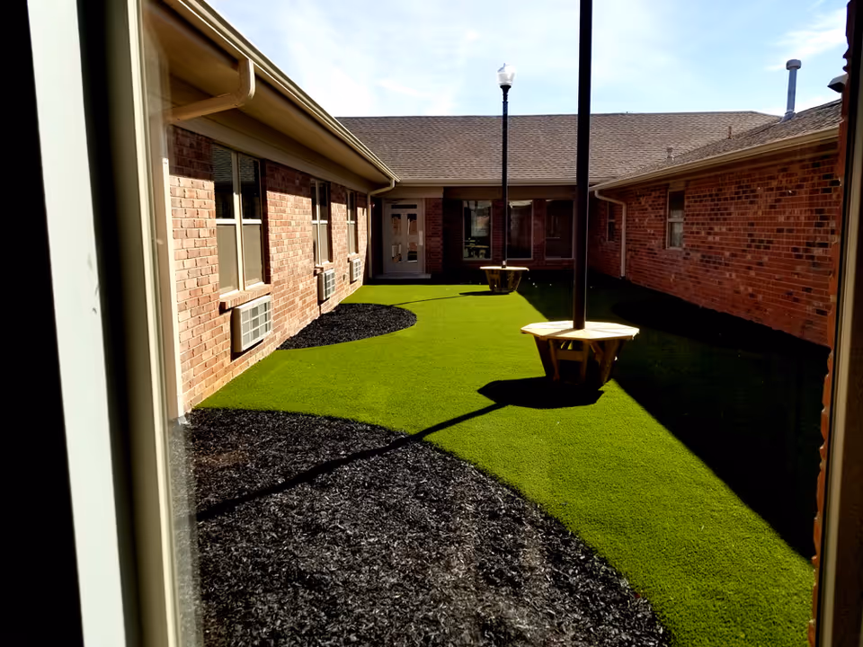 View through a window of a courtyard area with artificial green grass and black mulch patches, surrounded by brick walls of a building. There are two wooden benches around lamp posts in the courtyard.