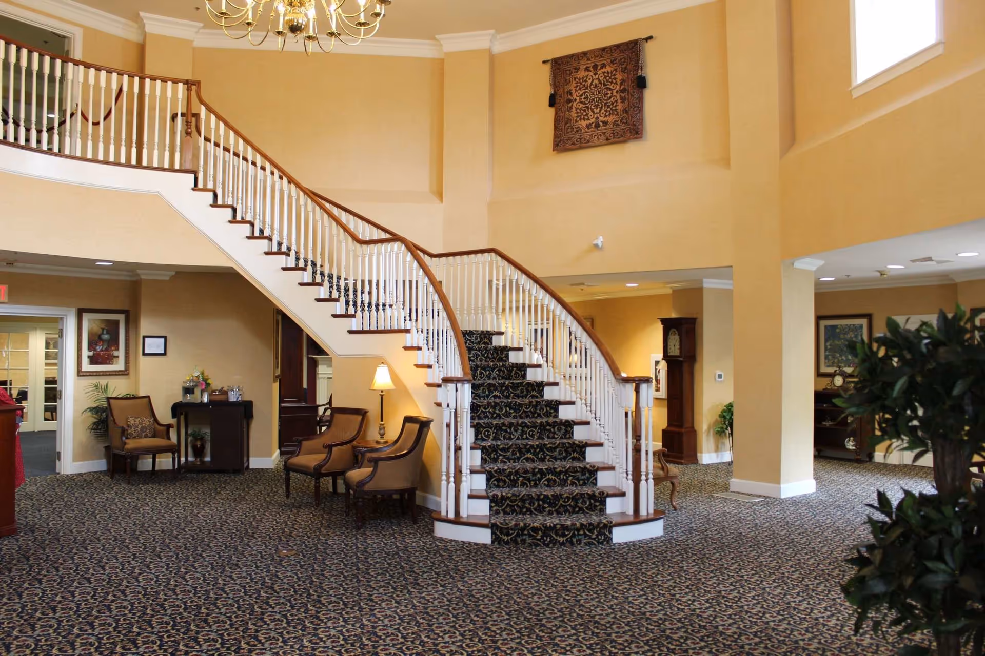 Interior of a senior living facility featuring a grand staircase with patterned carpet, beige walls, and a chandelier. There are several upholstered chairs arranged near the staircase, a small table with a lamp, framed artwork on the walls, and a large plant in the foreground. The space is well-lit with natural light coming from a window above.