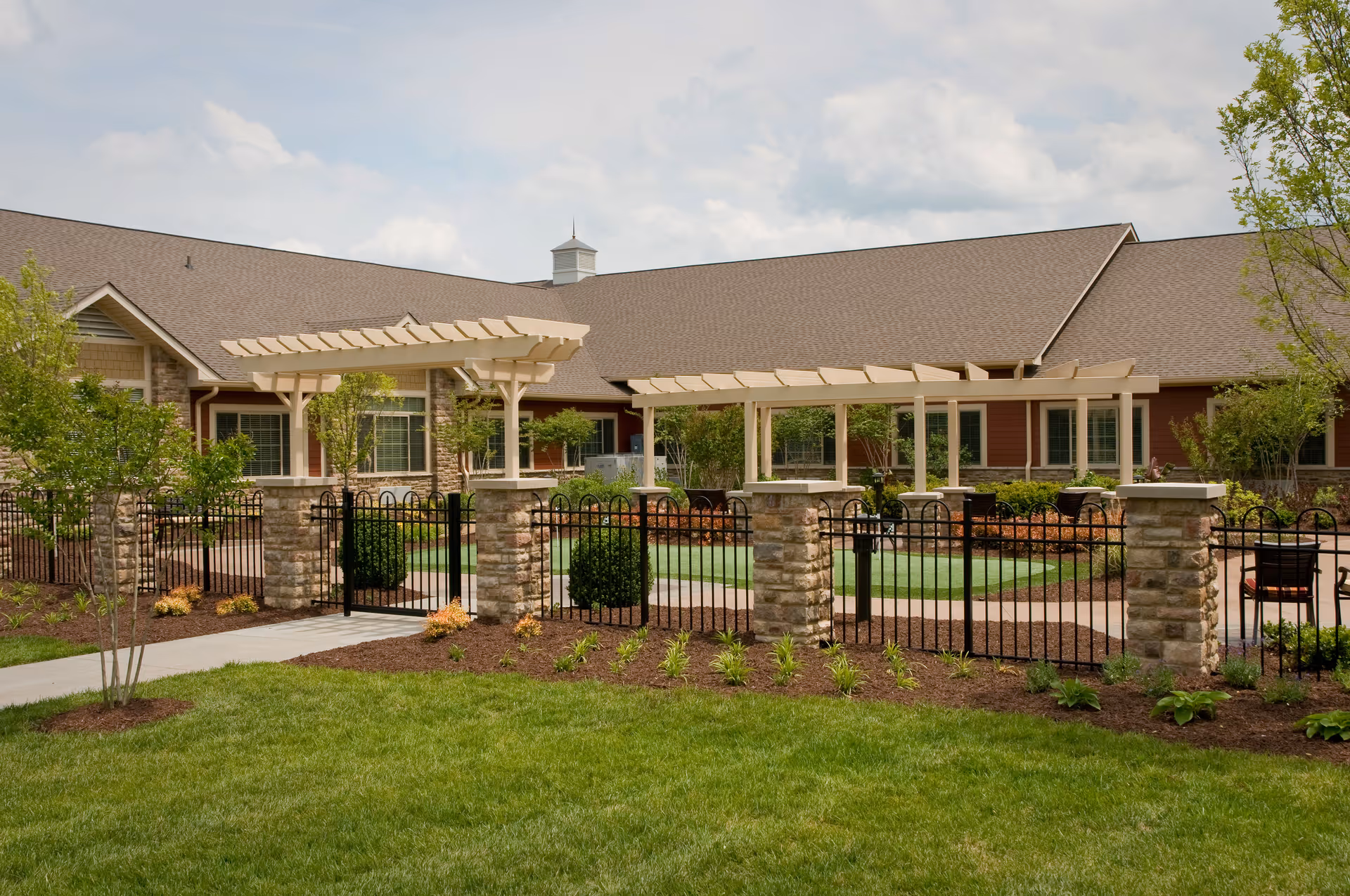 Exterior view of a single-story senior living facility with a fenced courtyard, pergolas, and landscaped lawn.