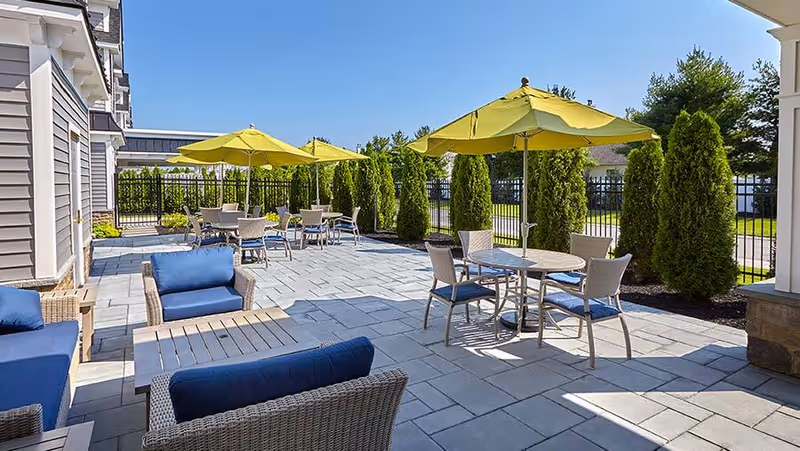 Outdoor patio area with several tables and chairs under yellow umbrellas, surrounded by tall green shrubs and a black metal fence, adjacent to a building with gray siding.
