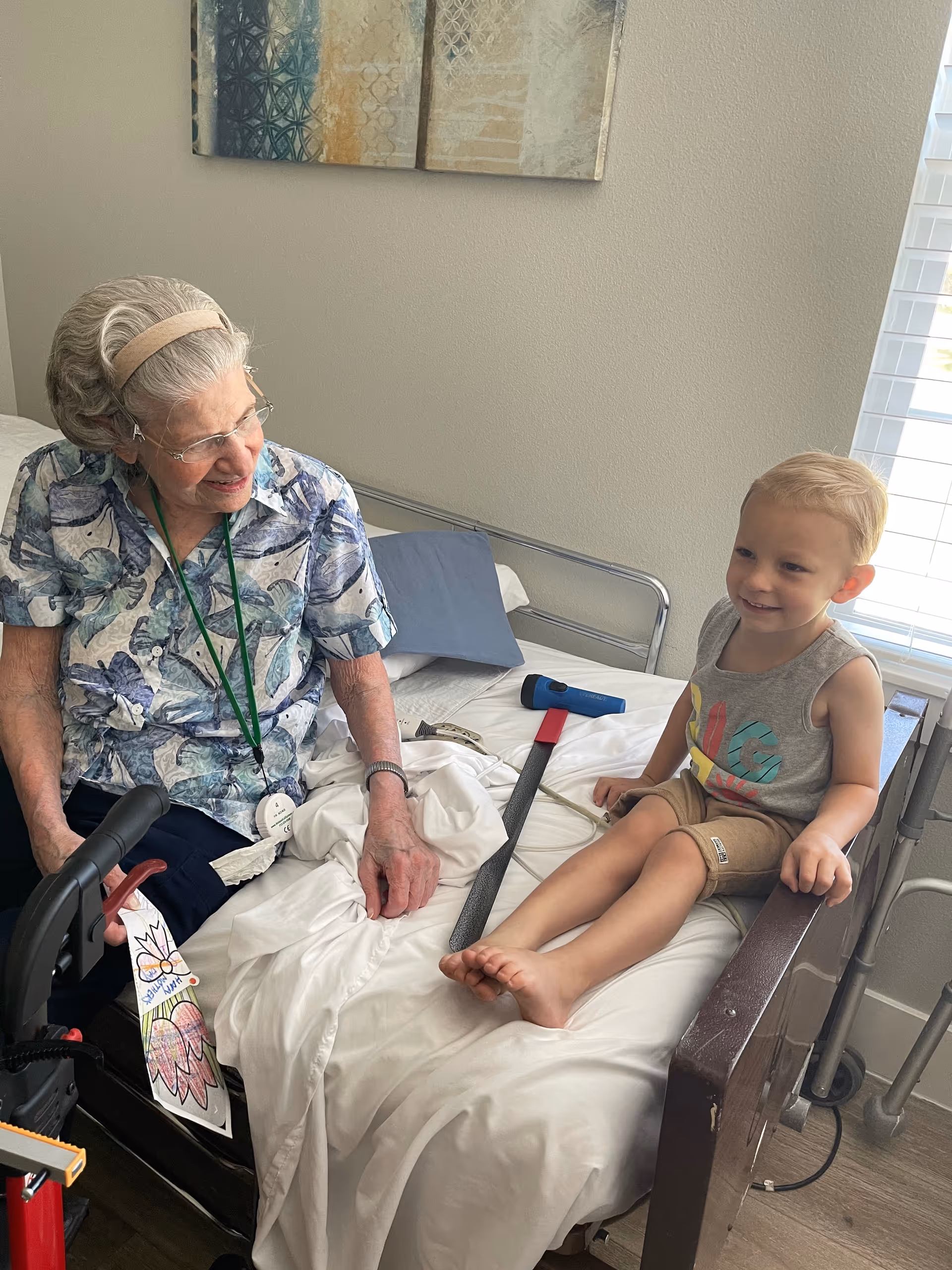 An elderly woman sitting on a bed in a senior care facility room, smiling and holding hands with a young boy who is sitting on the bed. The room has a window with blinds, a walker, and a colorful drawing attached to the walker.