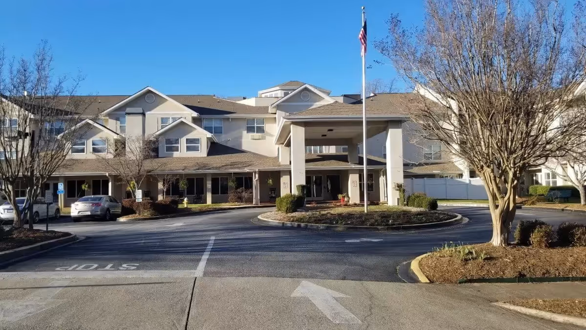 Front exterior view of TerraBella Greenville facility showing a multi-story building with a covered entrance, a flagpole with an American flag, several parked cars, and leafless trees under a clear blue sky.