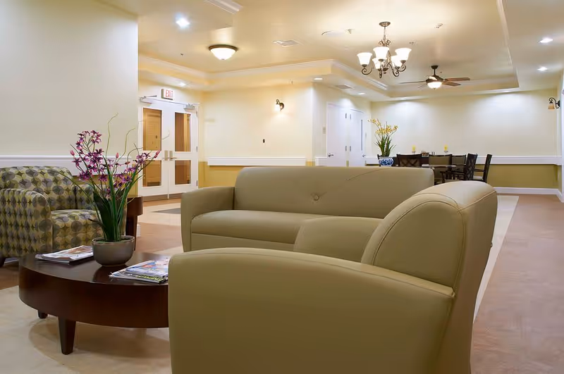 A well-lit assisted living facility common area with beige walls and ceiling lights. The room features a green leather sofa and armchair, a patterned armchair, a dark wooden coffee table with magazines and a potted plant with purple flowers. In the background, there is a dining table with chairs and another potted plant with yellow flowers. Double doors with glass panels are visible on the left side.