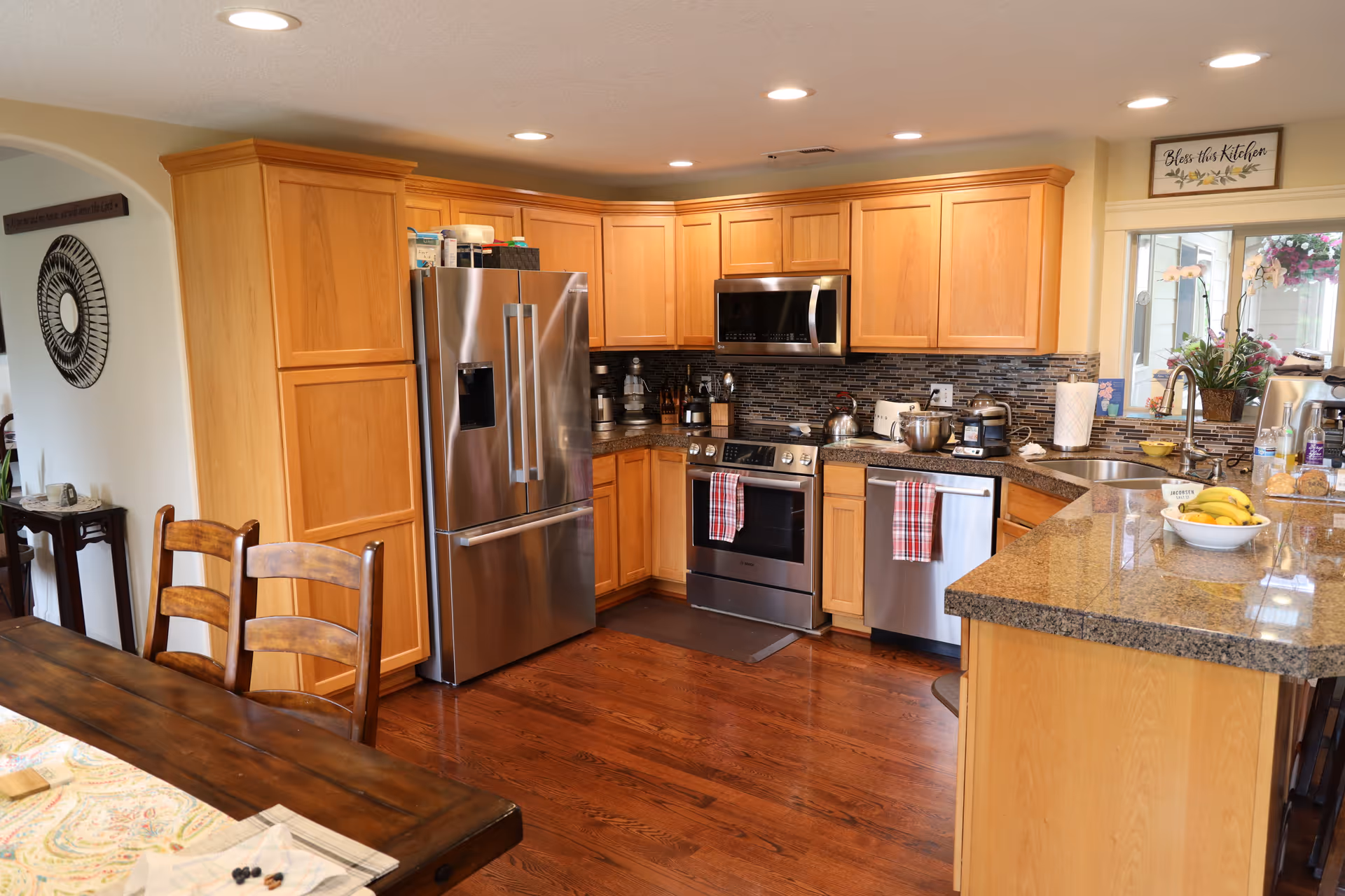 A modern kitchen with wooden cabinets, stainless steel refrigerator, oven, microwave, and dishwasher. The kitchen has a granite countertop with a bowl of bananas, a sink, and various kitchen appliances. There is a wooden dining table with chairs in the foreground and a sign above the window that reads 'Bless this Kitchen'.