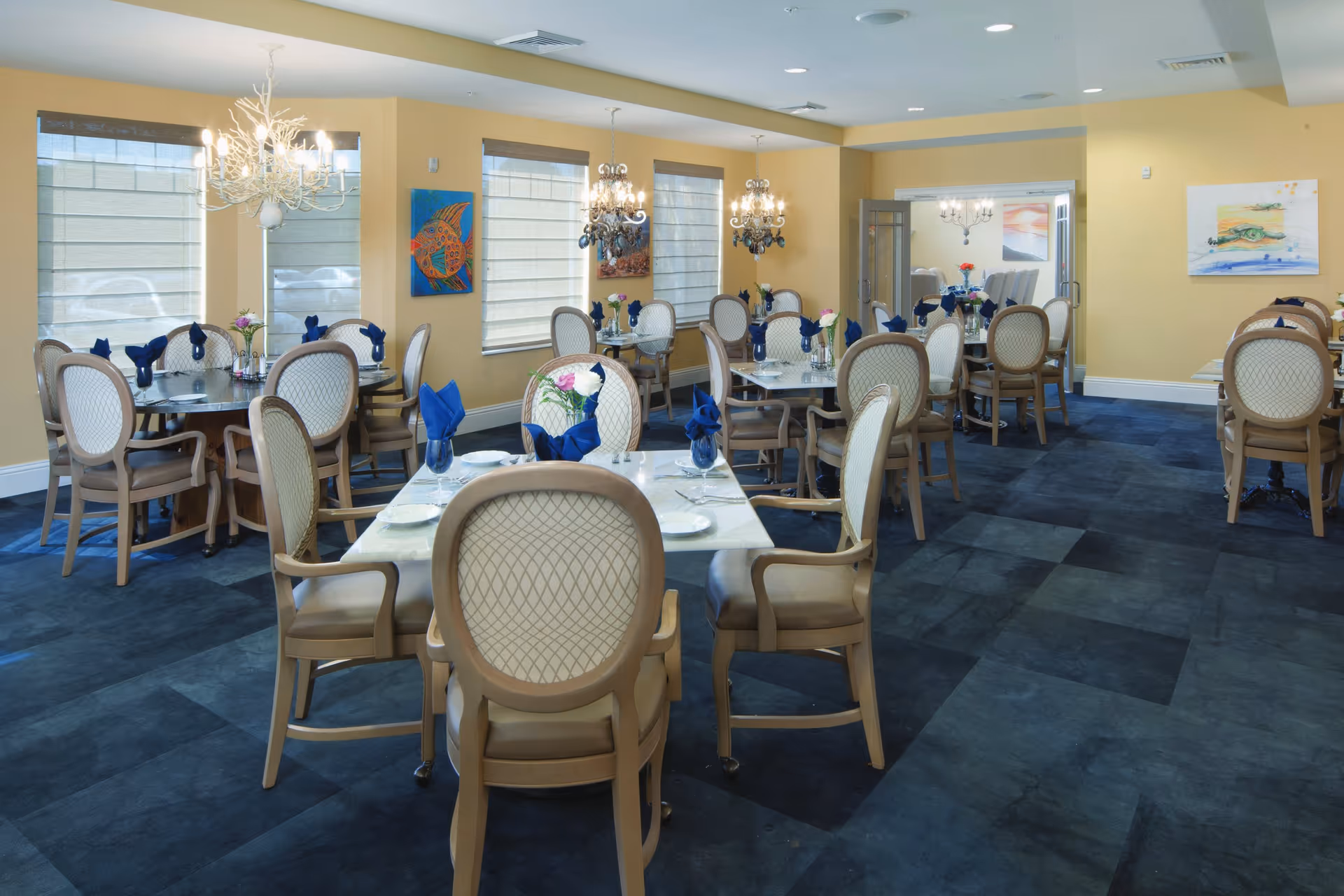 A bright dining room with multiple round and square tables set with blue napkins, chandeliers, and upholstered chairs.