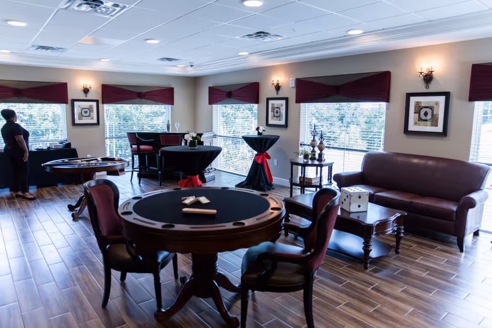 A well-lit common area with large windows covered by maroon valances, featuring a round card table with chairs, a brown leather sofa, a coffee table with a large decorative die, and two tall cocktail tables with black covers and red bows. A person stands near a table in the background, and the room has wood-look tile flooring and framed artwork on the walls.