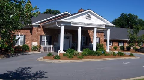 Front exterior view of a single-story brick building with white columns supporting a covered entrance. There are shrubs and small trees around the entrance, and a clear blue sky above.