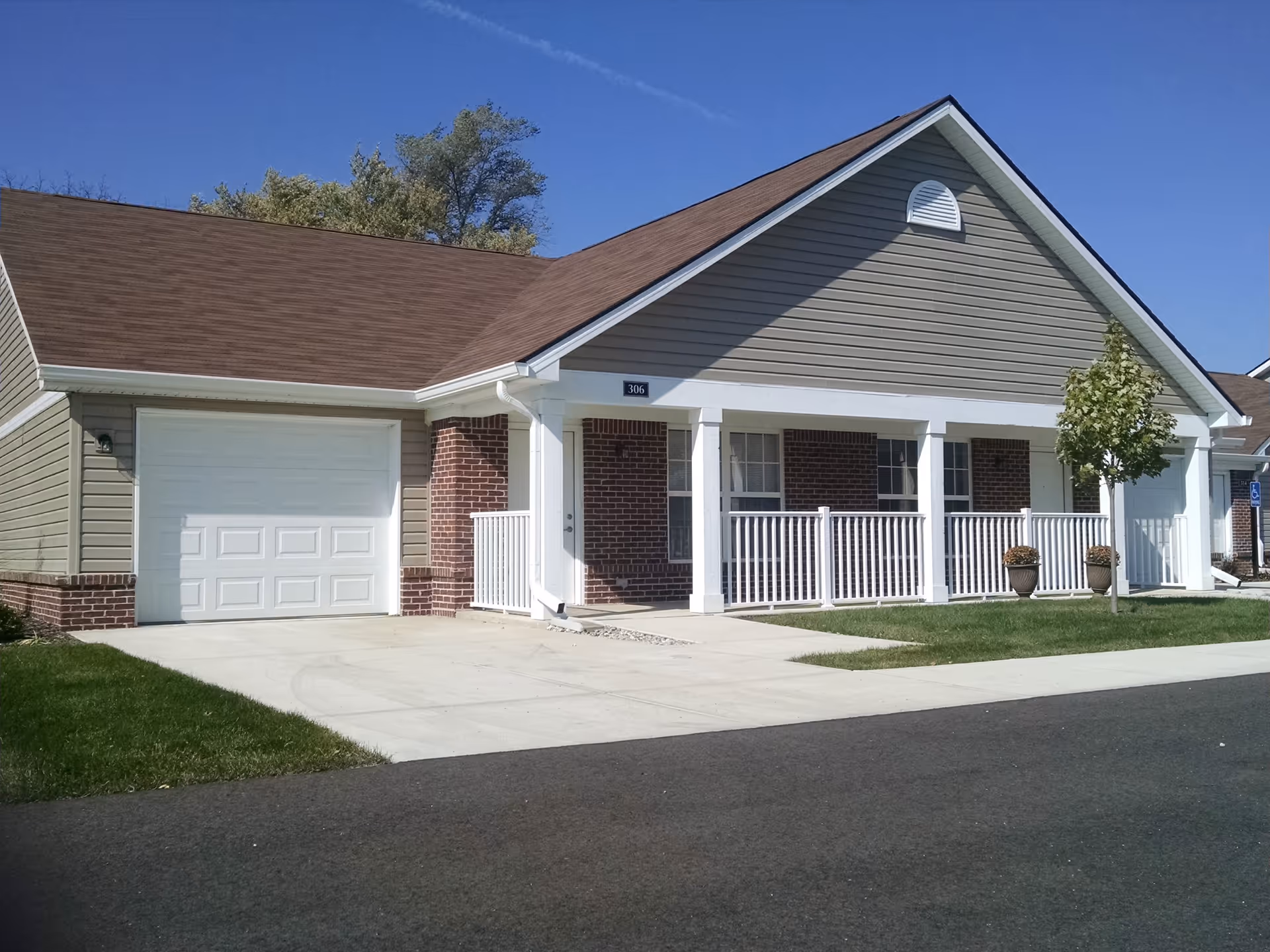 Exterior view of a single-story residential building with a brown shingled roof, beige siding, and brick accents. The building features a white garage door, a covered porch with white railings, two potted plants, a small tree, and a concrete driveway and walkway. The sky is clear and blue.