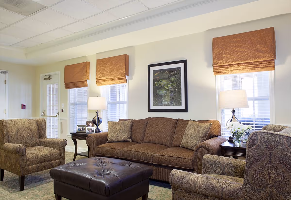 A cozy living room with a brown sofa, two patterned armchairs, and a large dark brown leather ottoman. The room has three windows with orange roman shades, two table lamps on side tables, framed artwork on the wall, and a vase with white flowers on one of the tables.