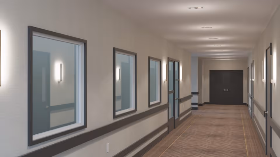 A long interior corridor in a senior living facility with wall-mounted lights, windows on the left, and doors along the hall.