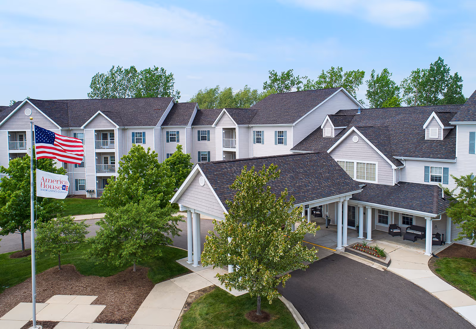 Exterior of American House Troy senior living campus with flag, trees, and covered entrance.