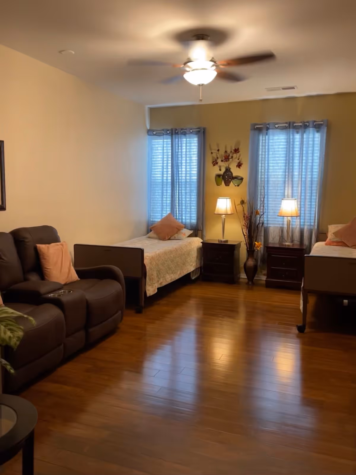 A cozy room with two single beds each with a pillow and floral bedspread, two nightstands with lamps between the beds, a brown leather recliner sofa with a pink pillow, wooden flooring, two windows with blue curtains, and a ceiling fan with a light.