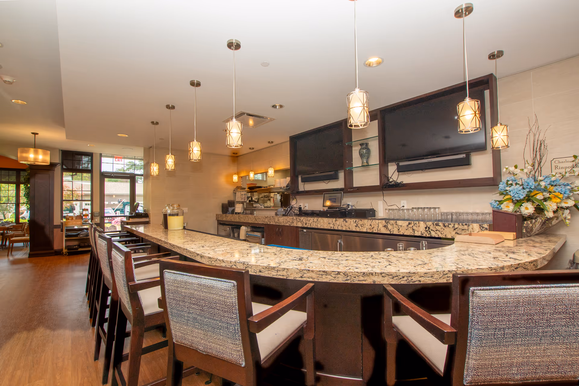 Interior view of a modern bar area in a senior living facility with a curved granite countertop, high chairs with cushioned seats, pendant lights hanging from the ceiling, two large flat-screen TVs mounted on the wall, and a flower arrangement on the counter. In the background, there is a dining area with tables and chairs near large windows.