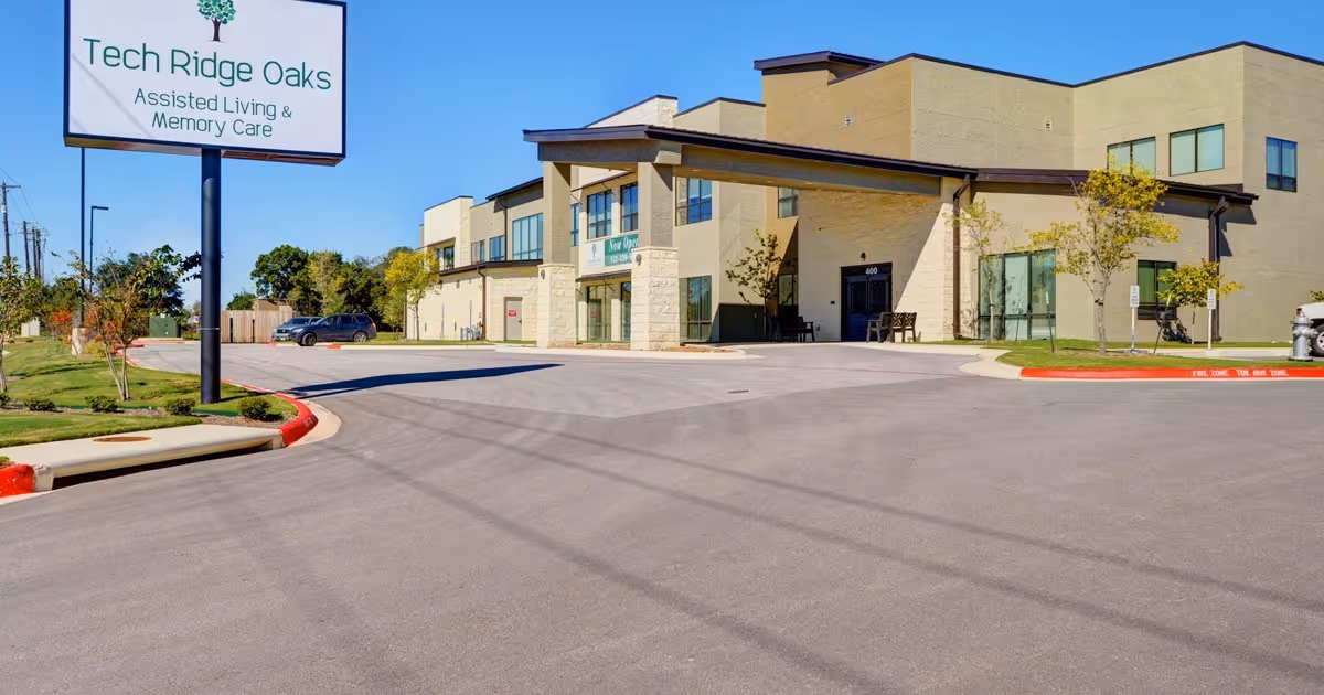 Exterior view of Tech Ridge Oaks Assisted Living & Memory Care facility with a large sign displaying the facility name. The building is modern with beige stone and large windows, surrounded by a paved driveway and some small trees and landscaping.