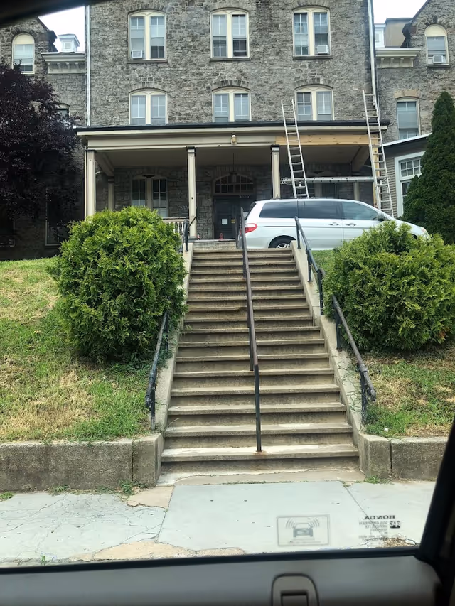 Front exterior of a stone multi-story building with concrete steps leading up to a covered porch, flanked by bushes and a white minivan parked by the entrance.