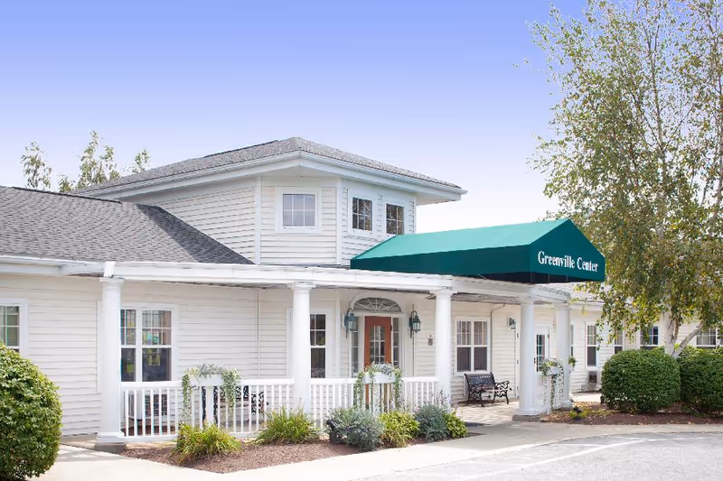 Exterior view of a white building with a green awning that reads 'Greenville Center'. The building has white columns supporting a porch area with benches and landscaping including bushes and trees around the entrance.