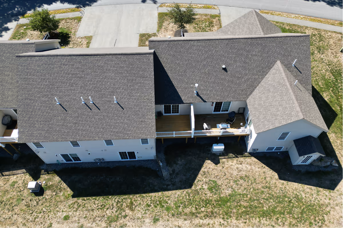 Aerial view of a residential building with a gray shingled roof and white exterior walls. The building has a small balcony with outdoor furniture and is surrounded by a patchy grass yard and paved driveway.