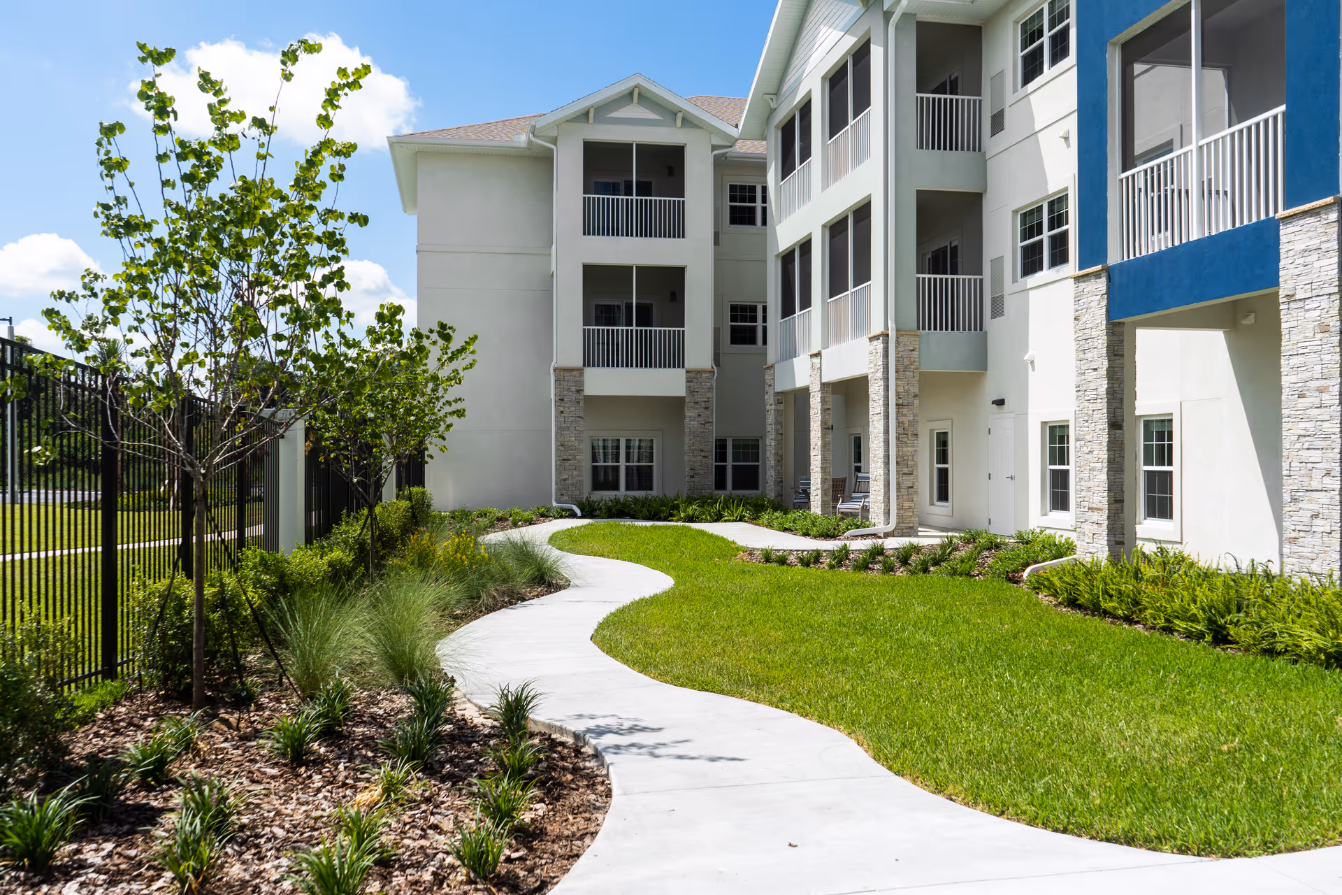 Curved concrete pathway surrounded by green grass, small trees, and plants leading to the entrance of a multi-story residential building with balconies and stone accents under a clear blue sky.