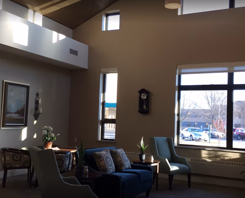 Sunlit common living area with chairs, a sofa, side tables, plants, a wall clock and large windows showing parked cars outside.