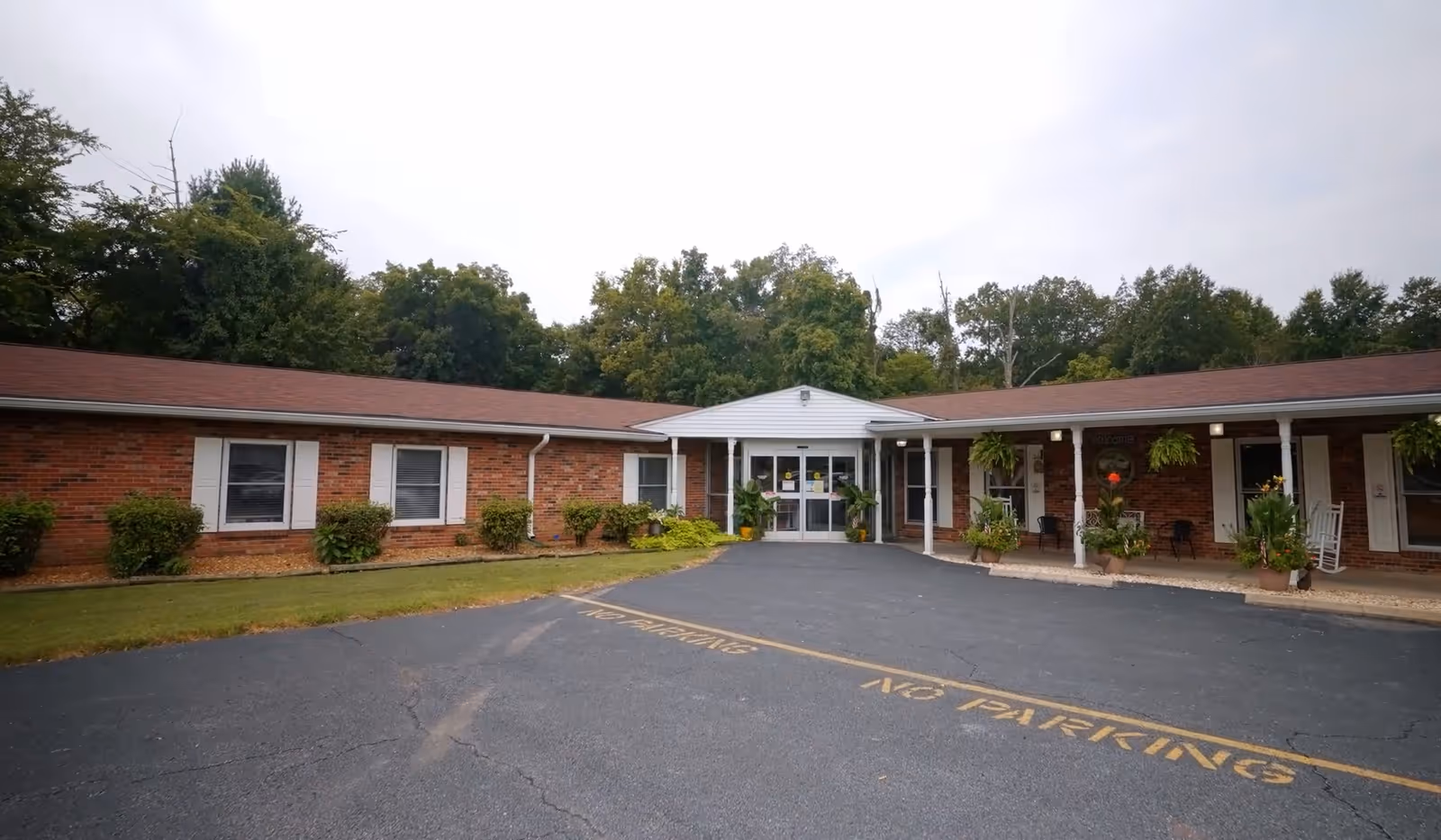 Front exterior view of a single-story brick building with white window shutters and a covered entrance. The building is surrounded by green shrubs and trees, with a paved driveway marked with 'NO PARKING' in yellow paint.
