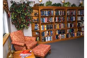 A cozy interior room featuring a large wooden bookshelf filled with books, a patterned armchair with a matching ottoman, a side table with magazines, and a tall leafy plant near a window.