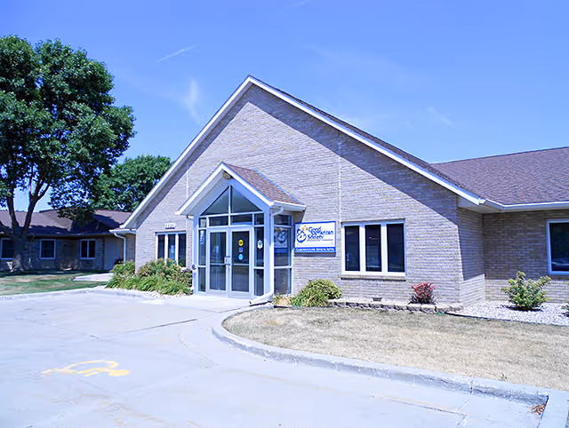 Exterior front view of a single-story brick building with a peaked roof and glass entrance doors. There is a sign next to the entrance that reads 'Good Samaritan Society'. The surrounding area includes a driveway, some landscaping with bushes, and a large tree on the left side under a clear blue sky.