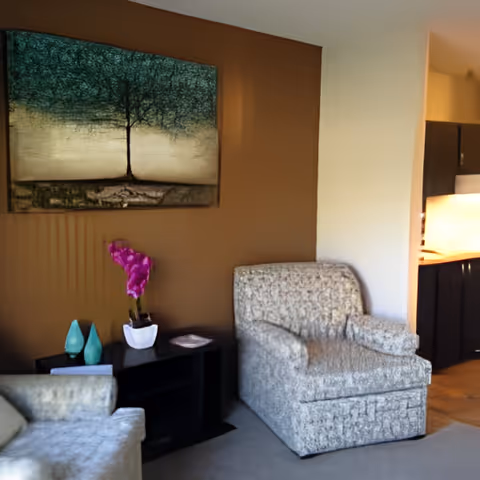A cozy living area with two patterned armchairs, a small black side table holding decorative items including a white pot with a pink orchid, and a large painting of a tree on a brown accent wall. To the right, there is a partial view of a kitchen with dark cabinets and a lit countertop.