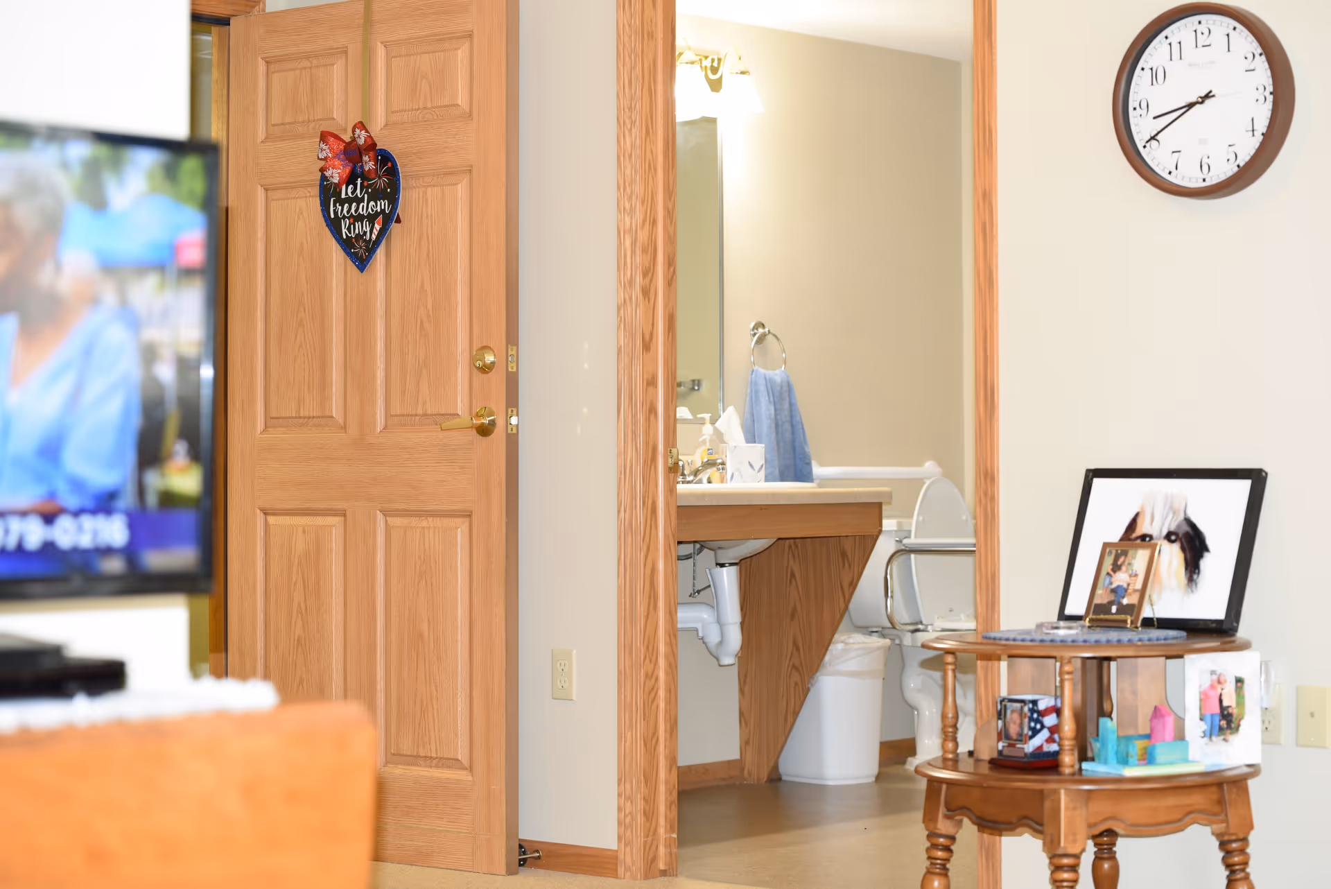 View of a partially open wooden door with a patriotic heart-shaped decoration that says 'Let freedom ring'. Through the doorway, a bathroom is visible with a sink, mirror, blue hand towel, and a toilet with grab bars. To the right, a wooden side table holds framed photos and a clock is mounted on the wall above.