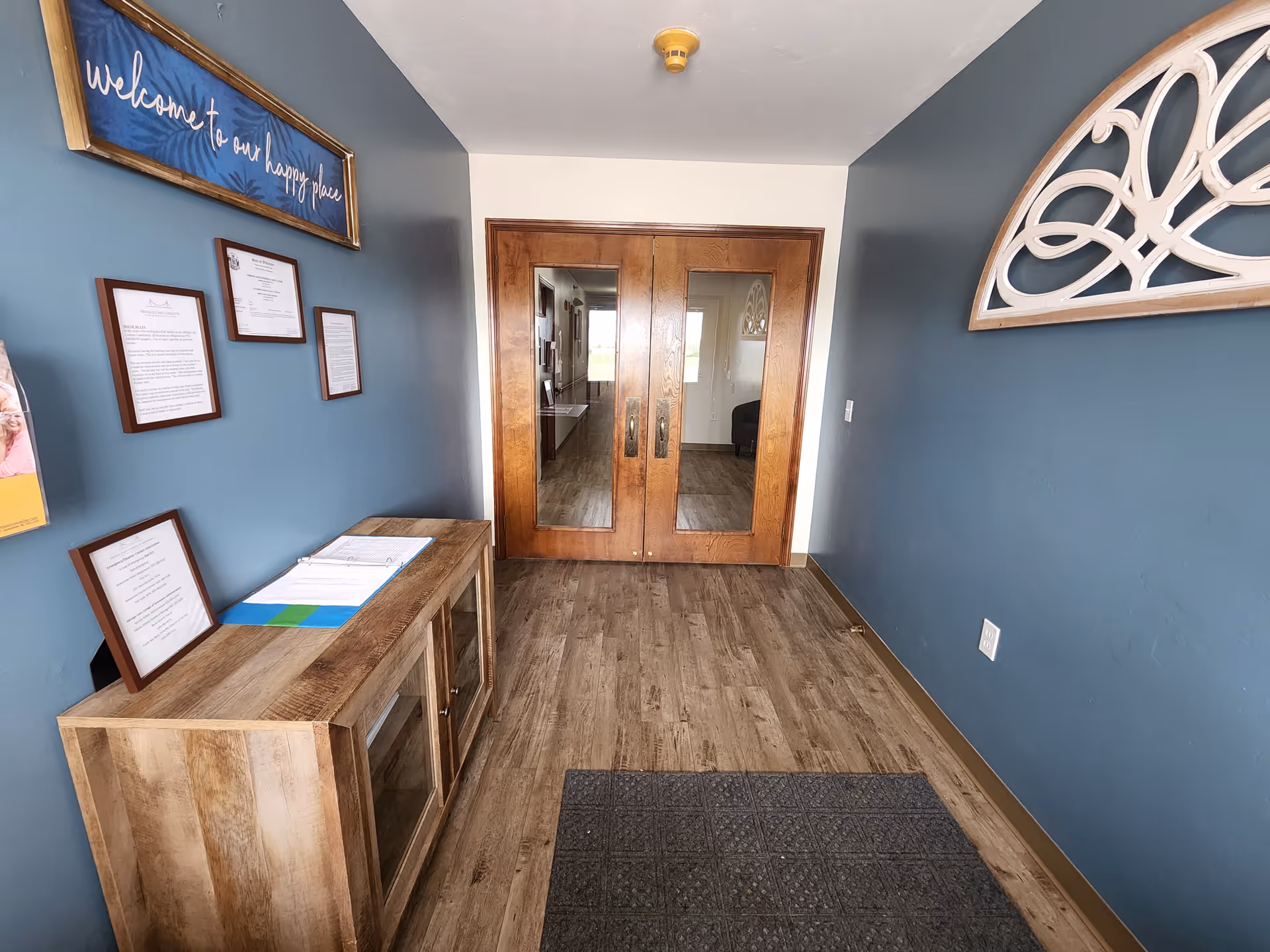 Entryway of a facility with wooden double doors featuring glass panels. The walls are painted blue and white, with a wooden cabinet on the left holding a binder and framed documents. Above the cabinet is a framed sign that reads 'welcome to our happy place.' A decorative wooden wall piece is on the right wall, and a gray floor mat is on the wooden floor.