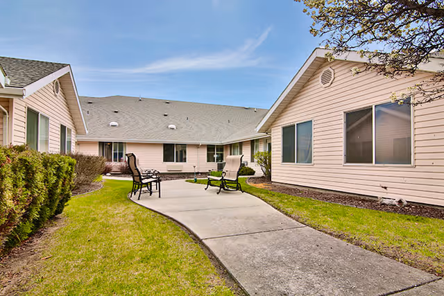 Outdoor courtyard area at Avamere at Hermiston with a concrete pathway, green grass, shrubs, and several chairs arranged for seating. The courtyard is surrounded by single-story beige buildings with windows under a blue sky with some clouds.