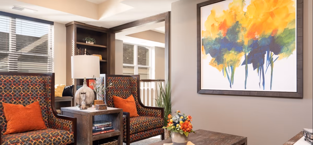 A cozy senior living common area with two patterned armchairs featuring orange cushions, a wooden side table with a lamp and decorative items, a colorful abstract painting on the wall, and a window with blinds letting in natural light.