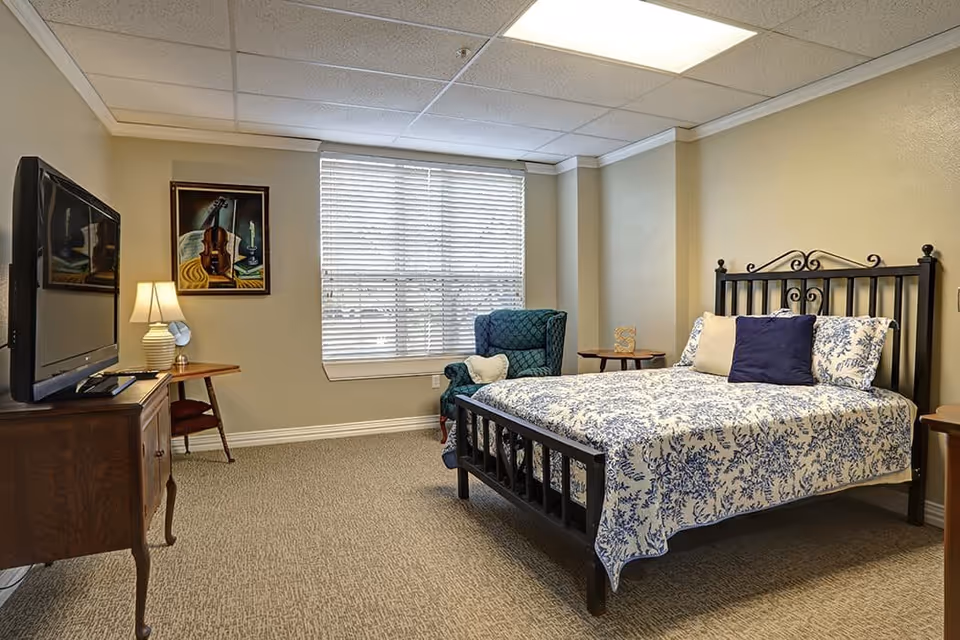 A cozy bedroom with a black metal bed frame, a bed covered with a white and blue floral patterned quilt, and three pillows. There is a green upholstered armchair with a heart-shaped pillow near a window with white blinds. A wooden TV stand with a flat-screen TV and a table lamp is on the left side of the room. A framed painting of a violin and sheet music hangs on the wall above a small wooden side table and chair.