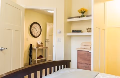 Bedroom interior with a bed footboard, built-in shelves holding towels and decor, and an open doorway revealing a hallway with a large wall clock and small table.