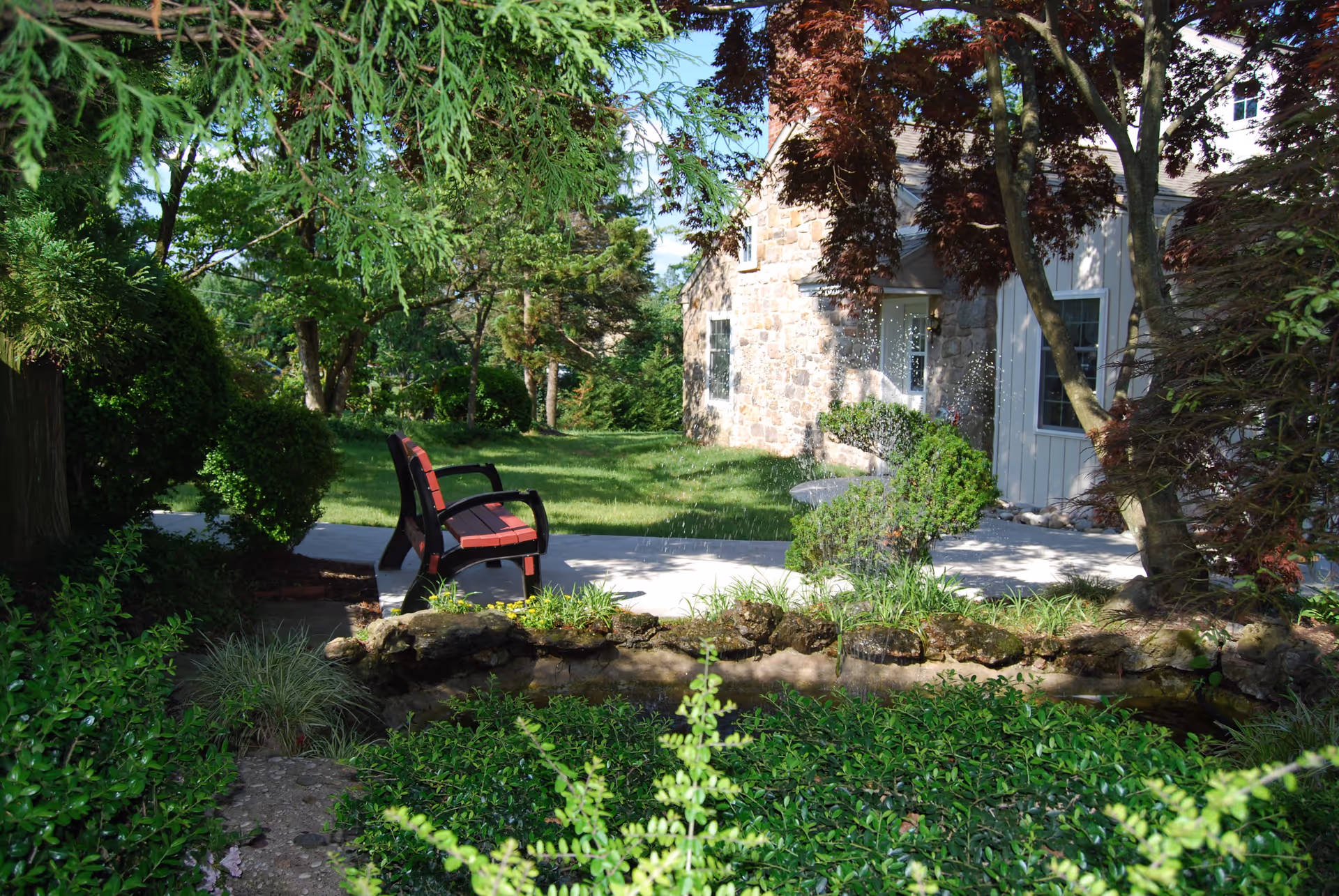 A peaceful outdoor garden area with lush green plants and trees surrounding a stone pathway. There is a red and black bench on the pathway, and a stone building with windows and a door is visible in the background.